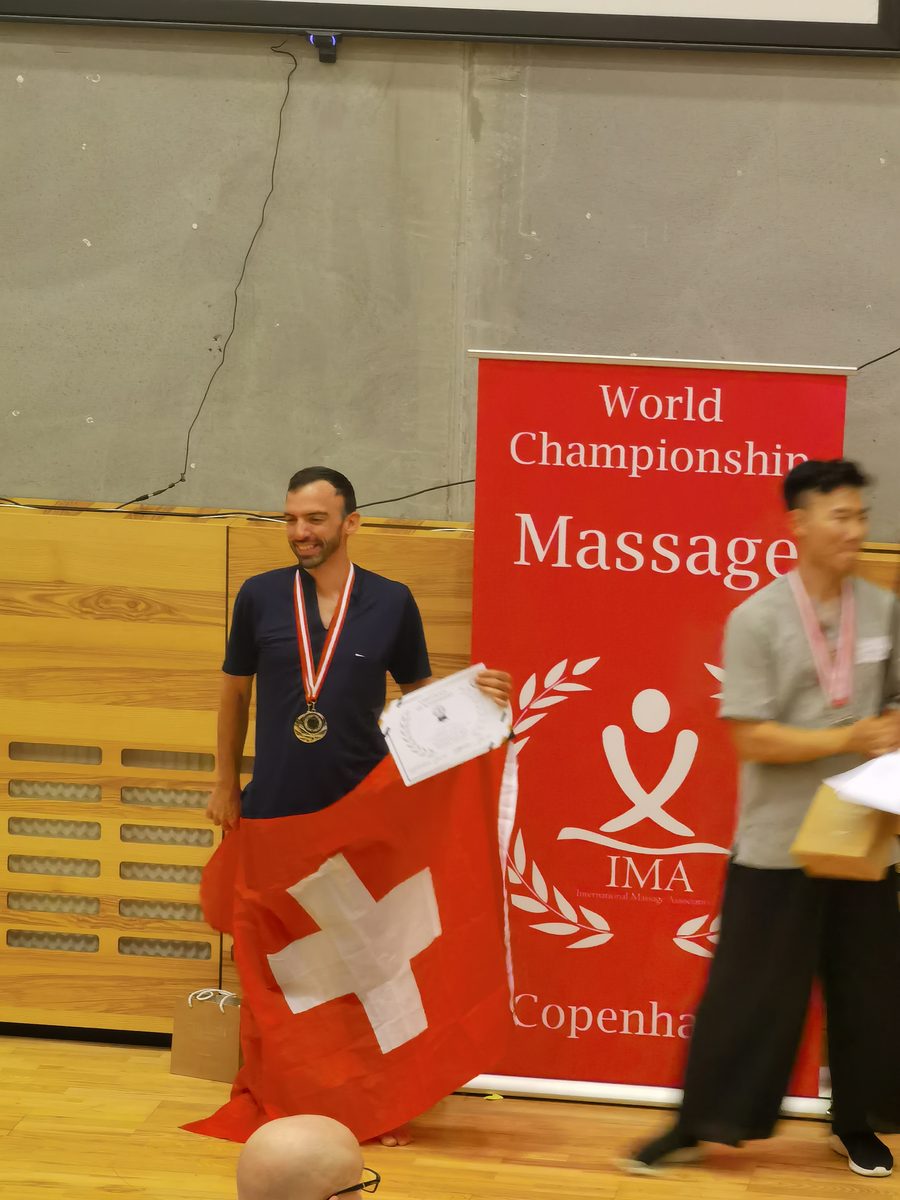 Laurent Lacour sur le podium du Championnat du Monde de Massage 2022 à Copenhague, médaille au cou, diplôme en main et drapeau suisse, devant la bannière de l'International Massage Association